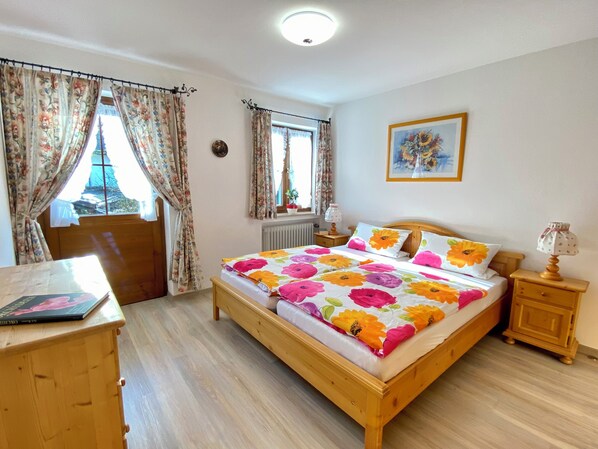 A bedroom with a wooden bed, floral bedding, a nightstand, a lamp, a window with curtains, and a painting on the wall.
