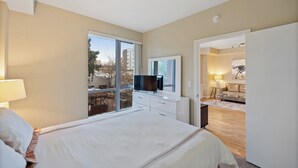 Un dormitorio moderno con una cama grande, un televisor sobre una cómoda y vistas al exterior a través de un amplio ventanal.
