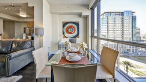 Un comedor moderno con una mesa de vidrio, sillas blancas y vistas a altos edificios a través de amplios ventanales.