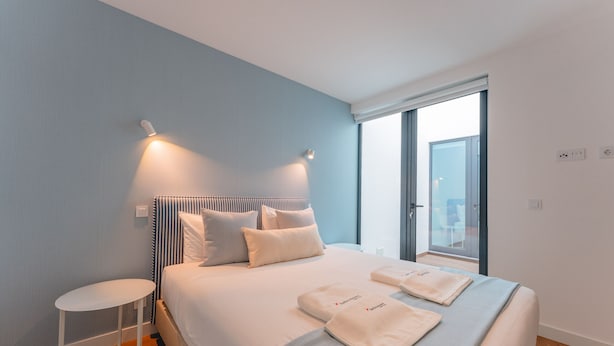 A modern hotel room with a large bed, two bedside tables, a small side table, and a large window offering a view of the city.