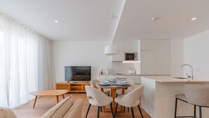 A modern living room with a sofa, a dining table with chairs, a TV, and a kitchen area with a microwave and sink.