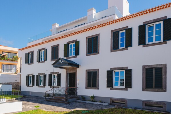 A white building with multiple windows and shutters, a covered entrance, and a small garden area.