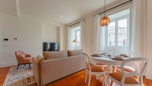 A modern living room with a sofa, dining table, and chairs. There is a TV on the wall and a window with curtains.