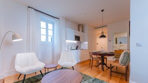 A modern living room with a dining area, white furniture, a round wooden table, and a kitchen in the background.