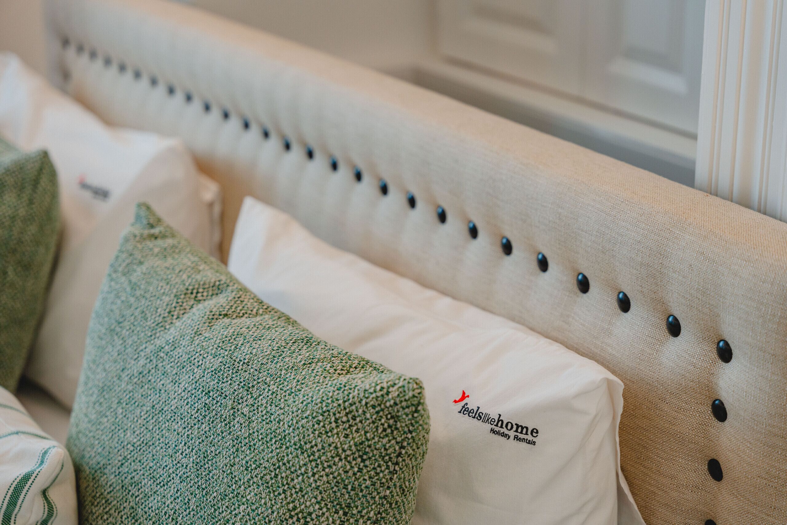 Una cama con cabecera capitonada, almohadas verdes y blancas, y una almohada con la marca 'feelslikehome'.