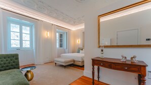 Ein Schlafzimmer mit großem Fenster, einem grünen Sessel, einer Holzkommode und einem weißen Bett.