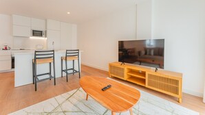 A modern living room with a wooden coffee table, two chairs, a TV, and a kitchen area with white cabinets and a microwave.