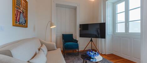 A living room with a beige sofa, a teal armchair, a flat-screen TV on a tripod stand, a floor lamp, and a large window with white curtains.