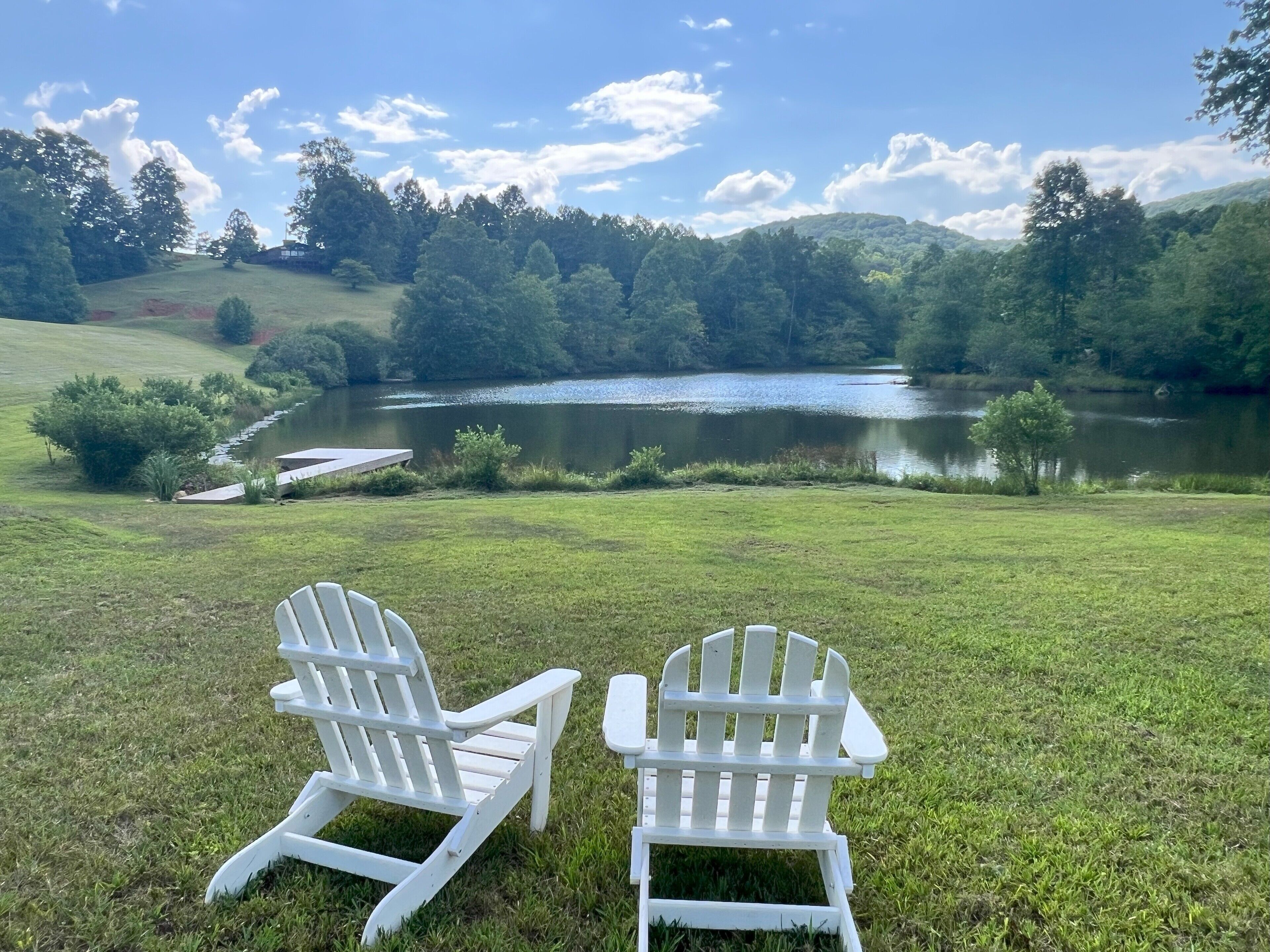 Dos sillas Adirondack blancas frente a un lago con un muelle y una zona verde.