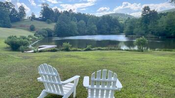 Two white Adirondack chairs facing a lake with a dock and a grassy area.