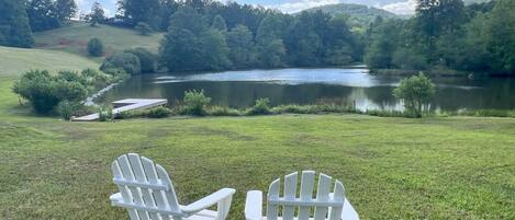 Dos sillas Adirondack blancas frente a un lago con un muelle y una zona verde.