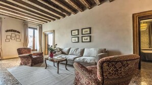 Un salon comprenant un canapé, deux fauteuils à motifs, une table basse et un tapis. Les murs sont ornés de tableaux encadrés et il y a une fenêtre avec des rideaux.
