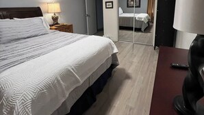 Habitación de hotel con una cama grande, un escritorio con lámpara, un espejo y una puerta que da a otra habitación.