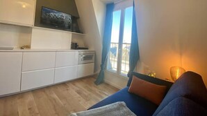 A modern living room with a sofa, a television mounted on a cabinet, a window with curtains, and a lamp. - Top floor Paris studio - view of rooftops and monuments  (Paris)