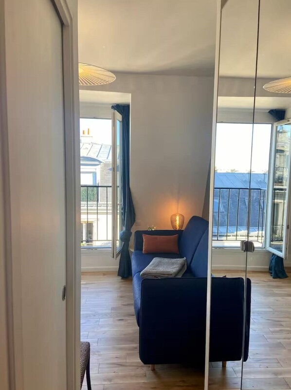 A modern living room with a dark blue sofa, a small table with a lamp, and large windows offering a view of the city. - Parisian Attic Studio—View of Rooftops and Landmarks   (Paris)