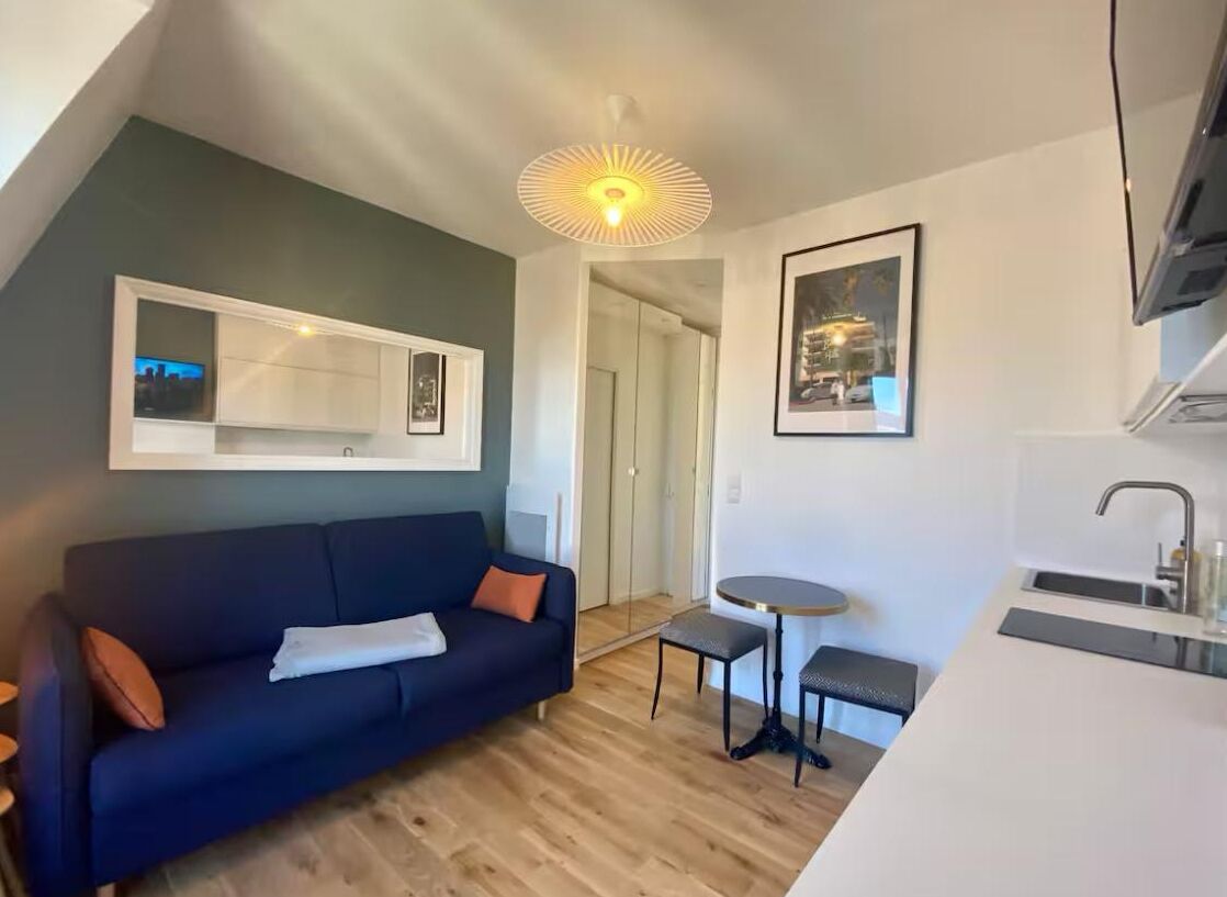 Um espaço compacto com um sofá azul escuro, uma mesa redonda pequena, cozinha planejada e um quadro na parede.