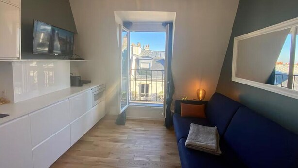 A modern kitchen with a built-in TV, a dining area with a blue sofa, and a view of the city through a large window. - Top floor Paris studio - view of rooftops and monuments  (Paris)