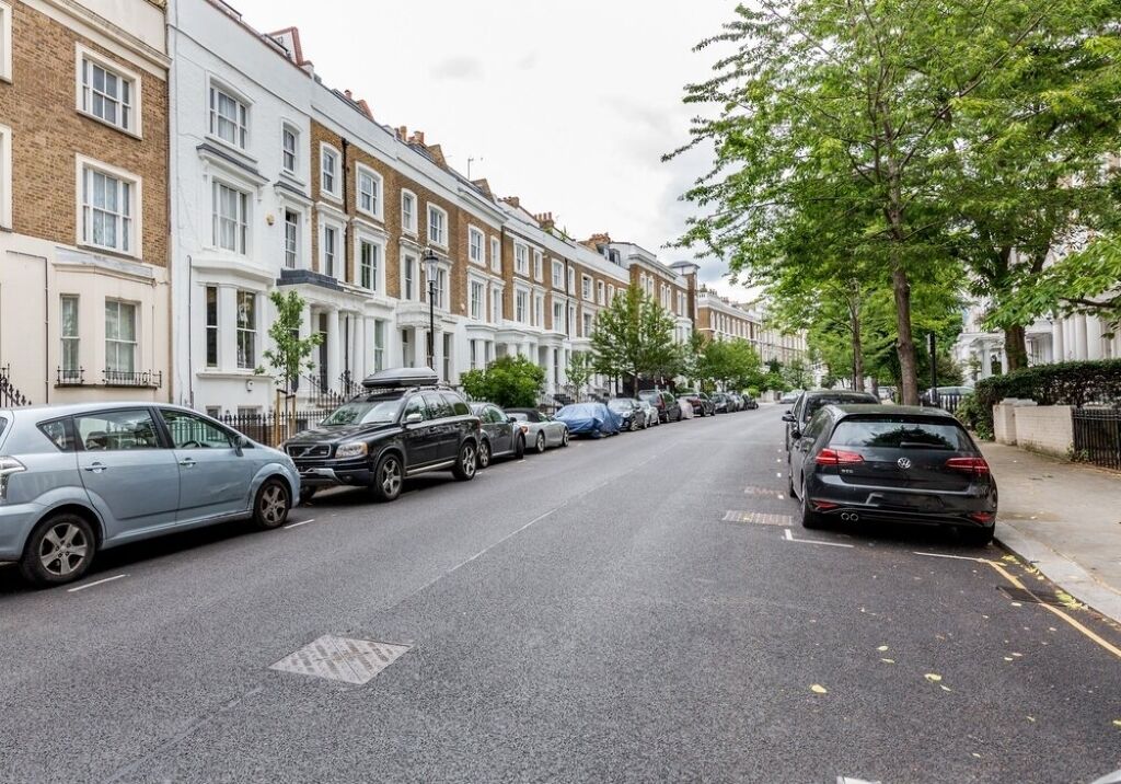 Eine Straße mit Reihenhäusern, parkenden Autos und Bäumen.