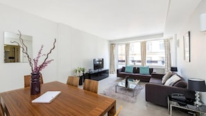 A modern living room with a wooden dining table, a sofa set, a TV, and a large window with curtains. - Wembley Empire Apartment - City Living (Wembley)