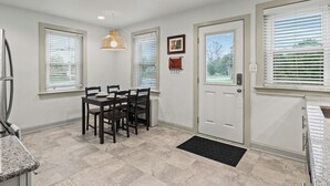 Uma cozinha com mesa de jantar, cadeiras e geladeira. Há janelas com persianas, uma porta e um lustre sobre a mesa.