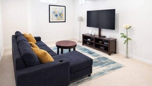 Un salon avec un canapé bleu foncé, une table basse ronde, un meuble TV sur lequel est posée une télévision à écran plat, une lampe sur pied et un vase contenant une seule fleur blanche.
