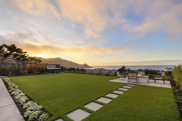 A well-maintained backyard with a grassy lawn, a fire pit, and a seating area overlooking a scenic ocean view.