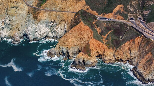 A coastal road with a bridge and tunnels, surrounded by rocky cliffs and ocean waves.