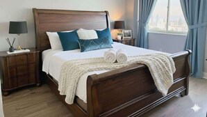 Una cama bien hecha con edredón blanco y almohadas azules, cabecera de madera y vista a un paisaje urbano a través de una ventana con cortinas azules.