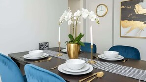 Una mesa de comedor dispuesta con platos blancos, fuentes y cubiertos dorados, rodeada de sillas azules.