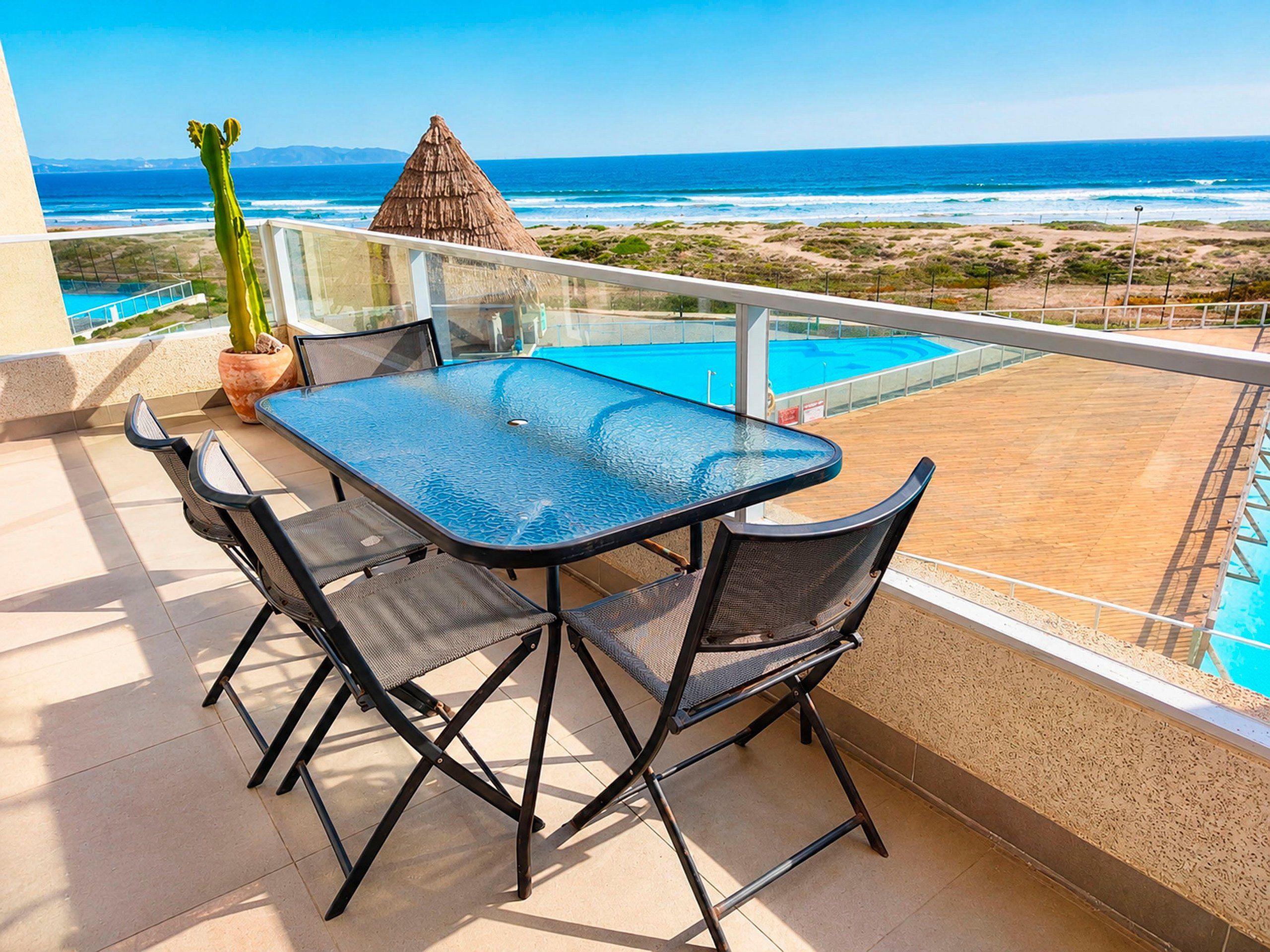 Une table à plateau en verre, quatre chaises, une plante en pot et une structure à toit de chaume sur un balcon donnant sur une piscine et une plage.
