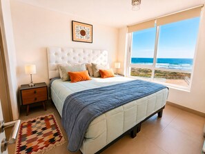 Een slaapkamer met een bed, twee nachtkastjes, een vloerkleed en een raam met uitzicht op de oceaan.