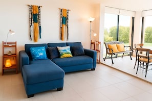 A modern living room with a blue sofa, a wooden side table, a floor lamp, and a view of the outdoors through large windows.