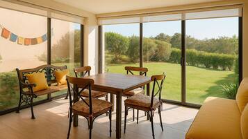 Une terrasse ensoleillée avec une table et des chaises en bois, un banc rembourré et une vue sur un paysage verdoyant, visible à travers de grandes fenêtres.