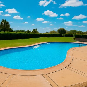 Piscina azul límpida com borda curva, rodeada por um deck pavimentado e um gramado bem cuidado.