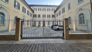 Une cour dotée d’une entrée fermée par un portail, d’un chemin pavé et d’un bâtiment avec de nombreuses fenêtres et des arcades.