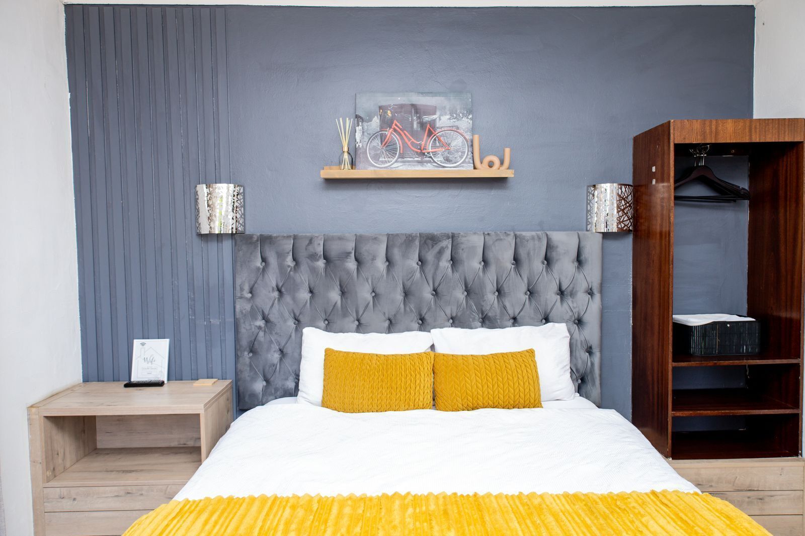 A bedroom with a tufted headboard, a bed with white and yellow bedding, a wooden nightstand, a wall-mounted shelf with a bicycle painting, and a wardrobe with hanging space.