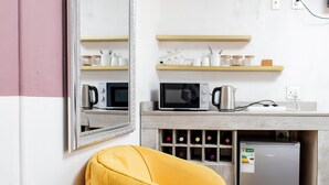 Une petite cuisine équipée d’un four à micro-ondes, d’une machine à café et d’un réfrigérateur à vin. Des étagères au-dessus du comptoir servent à ranger des bocaux et des verres. Une chaise jaune se trouve au premier plan.