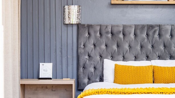 Une chambre à coucher avec une tête de lit capitonnée, un lit recouvert d’une couvre-lit blanche et jaune, une petite table de chevet avec une lampe, et une étagère murale ornée d’un vélo et d’objets décoratifs.