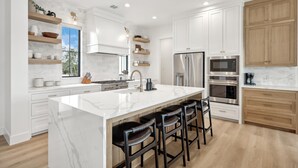 Una cocina moderna con armarios blancos, una isla grande con taburetes, electrodomésticos integrados y estantes de madera.