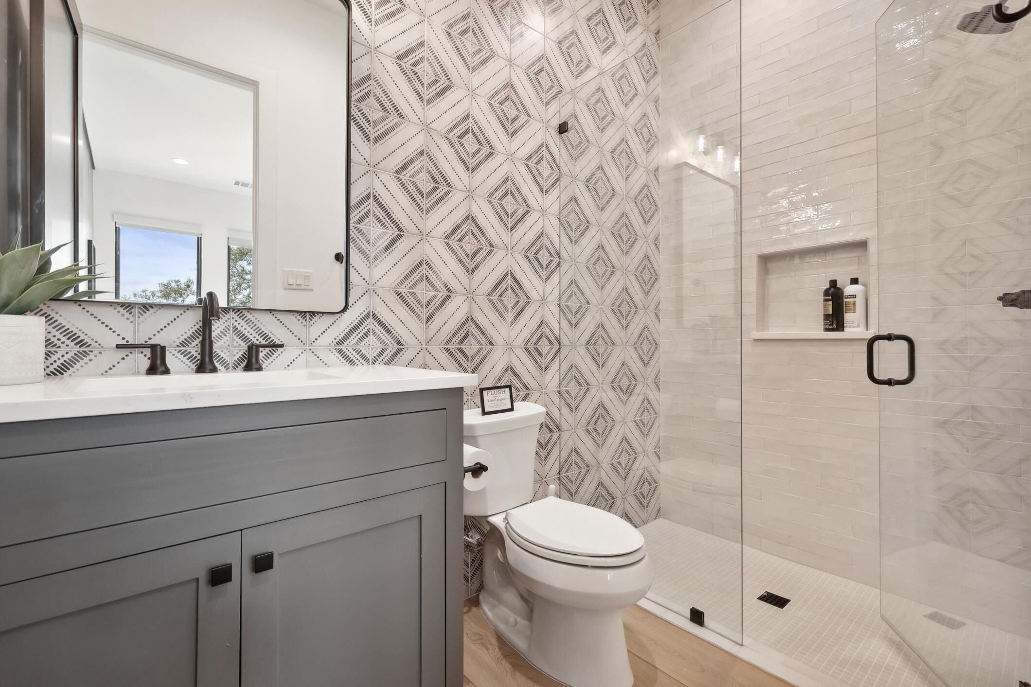 A modern bathroom with a geometric patterned wall, a white toilet, a glass-enclosed shower, and a vanity with a mirror.