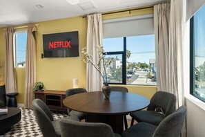 Een moderne kamer met een ronde tafel, stoelen, een televisie met 'NETFLIX', een raam met gordijnen en uitzicht op buiten.
