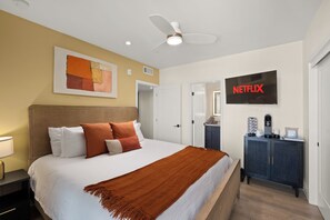 Een hotelkamer met een bed, een plafondventilator, een aan de muur bevestigde televisie waarop het Netflix-logo te zien is, en een koffieapparaat op een kast.