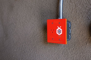 Een rode elektriciteitskast bevestigd aan een gestructureerde muur, met een QR-code en een symbool dat een brandalarm aanduidt.