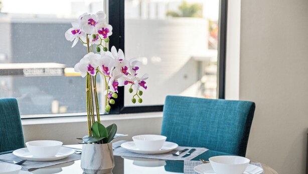 Een eettafel gedekt met witte kommen, borden en bestek, een vaas met witte orchideeën, en teal-kleurige stoelen.