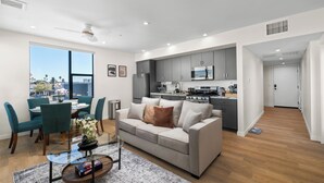 Een moderne woonkamer met een grijze bank, blauwgroene fauteuils en een glazen salontafel. Op de achtergrond is een keuken met grijze kastjes en een magnetron zichtbaar.