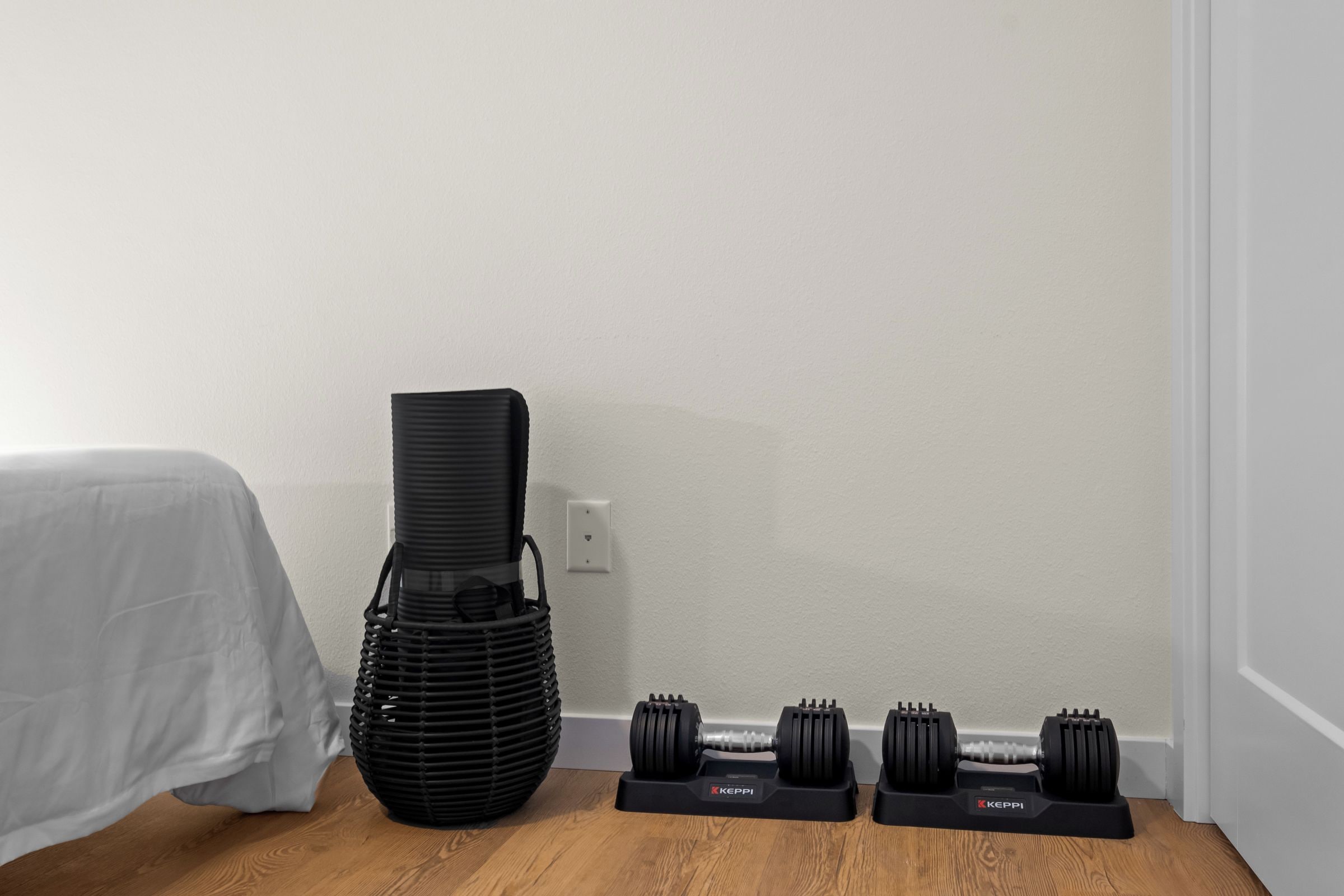 A corner of a room with a bed, a black exercise mat, and a set of dumbbells on a stand.