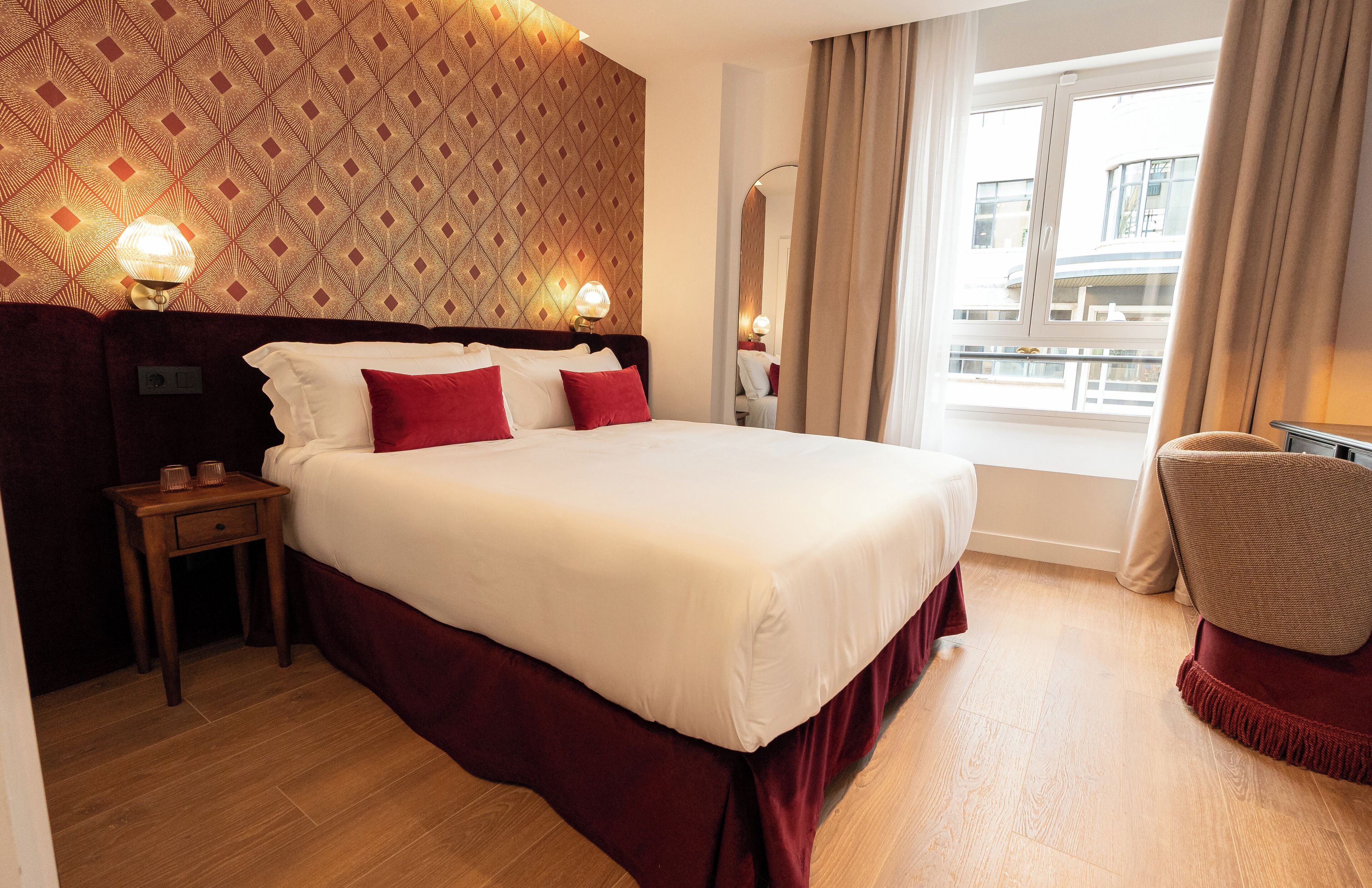 Een hotelkamer met een groot bed, twee bedlampjes, een houten nachtkastje, een stoel en een raam met gordijnen.