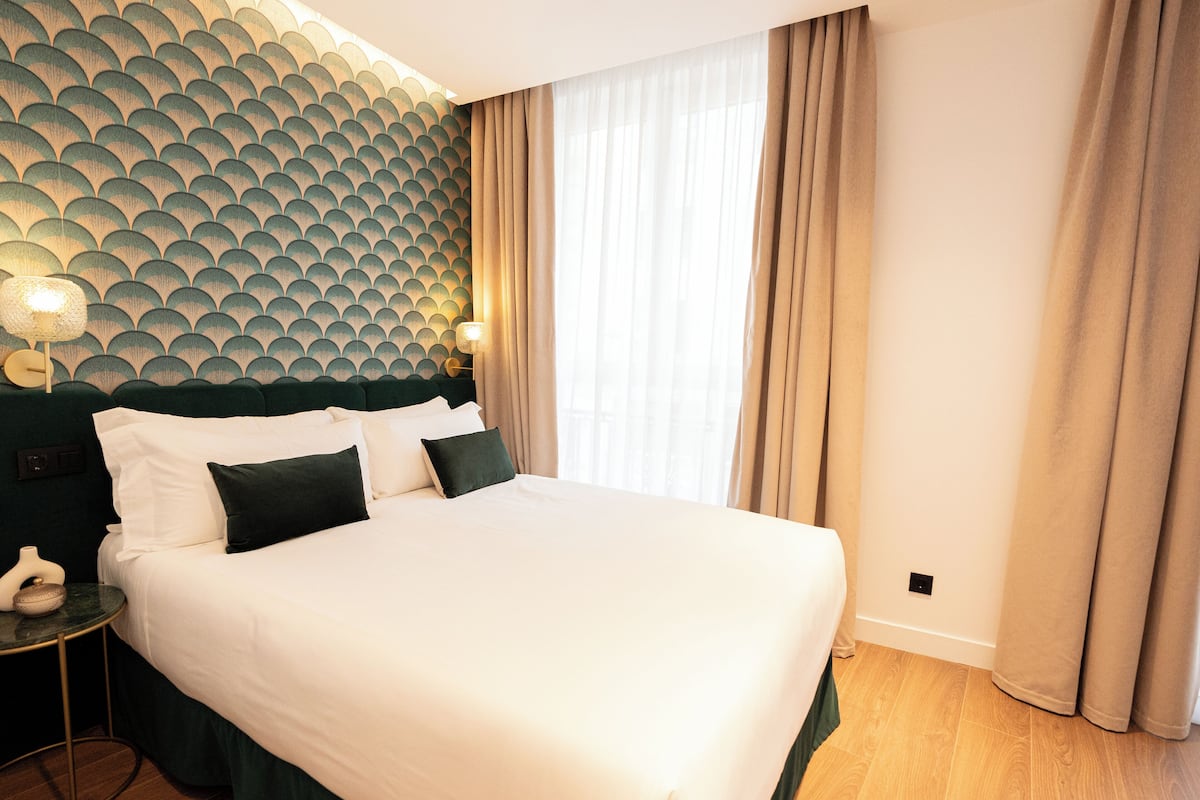A neatly made bed with white linens and dark green bedding, a round side table with a lamp, and a wall with a geometric pattern.