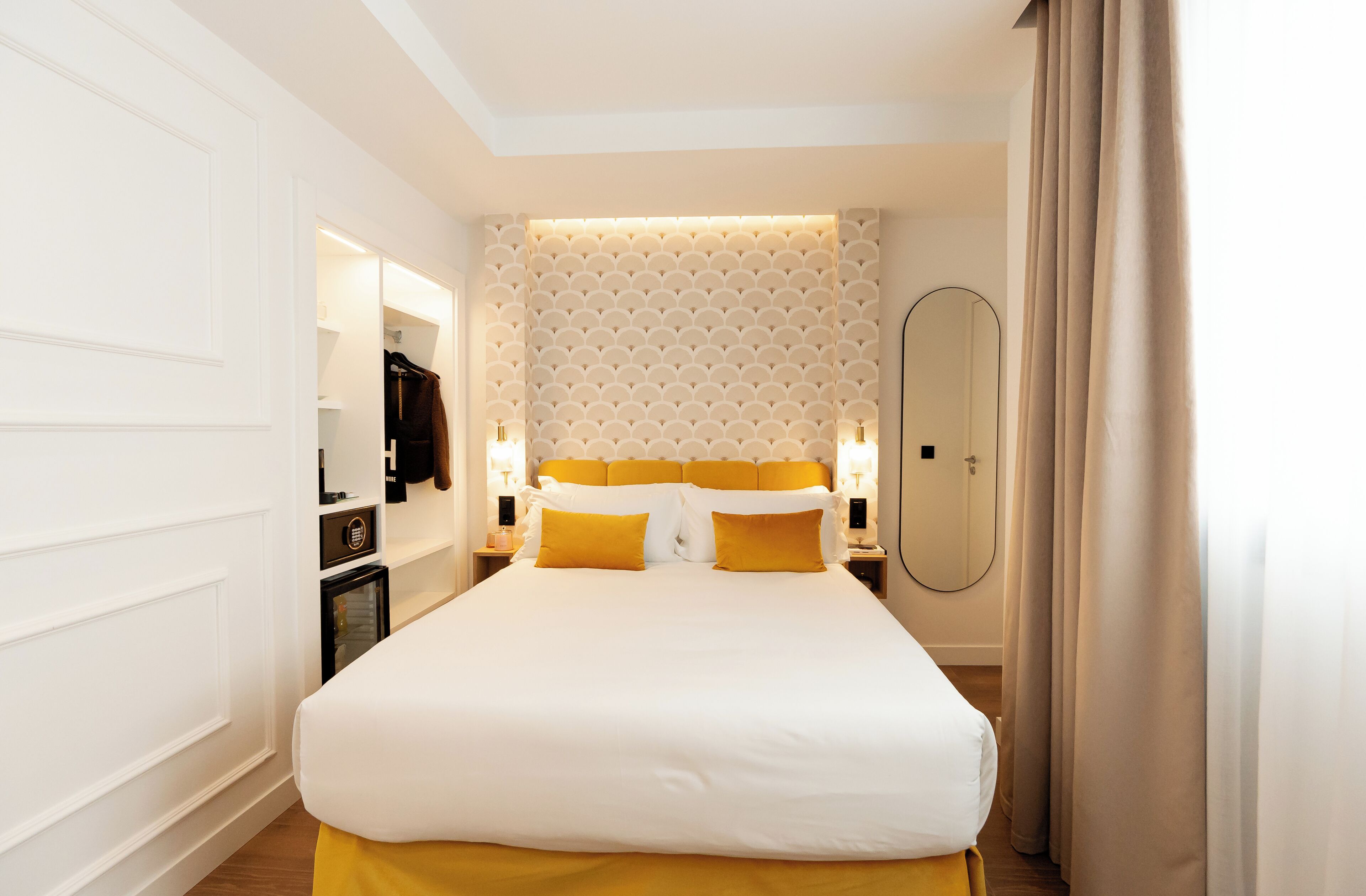 Una habitación de hotel con una cama grande, un armario, un espejo y una mesita de noche con lámpara.