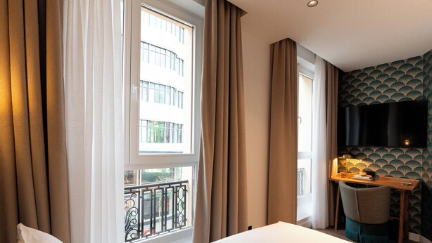 Une chambre d’hôtel comprenant un lit, un bureau avec une chaise, un téléviseur fixé au mur et une fenêtre avec des rideaux.